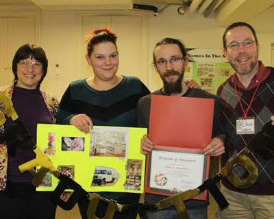 4 people together. one is holding a poster of pictures. the other is holding up a certificate