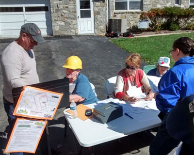 people at a table outside on a driveway. one person has a safety helmet on.